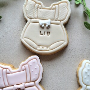 May include: Three decorated sugar cookies shaped like baby onesies. The top cookie is beige with a white bow and the name "LIU" written on it. The middle cookie is pink with a white bow and the name "ANNA" written on it. The bottom cookie is white with a white bow.