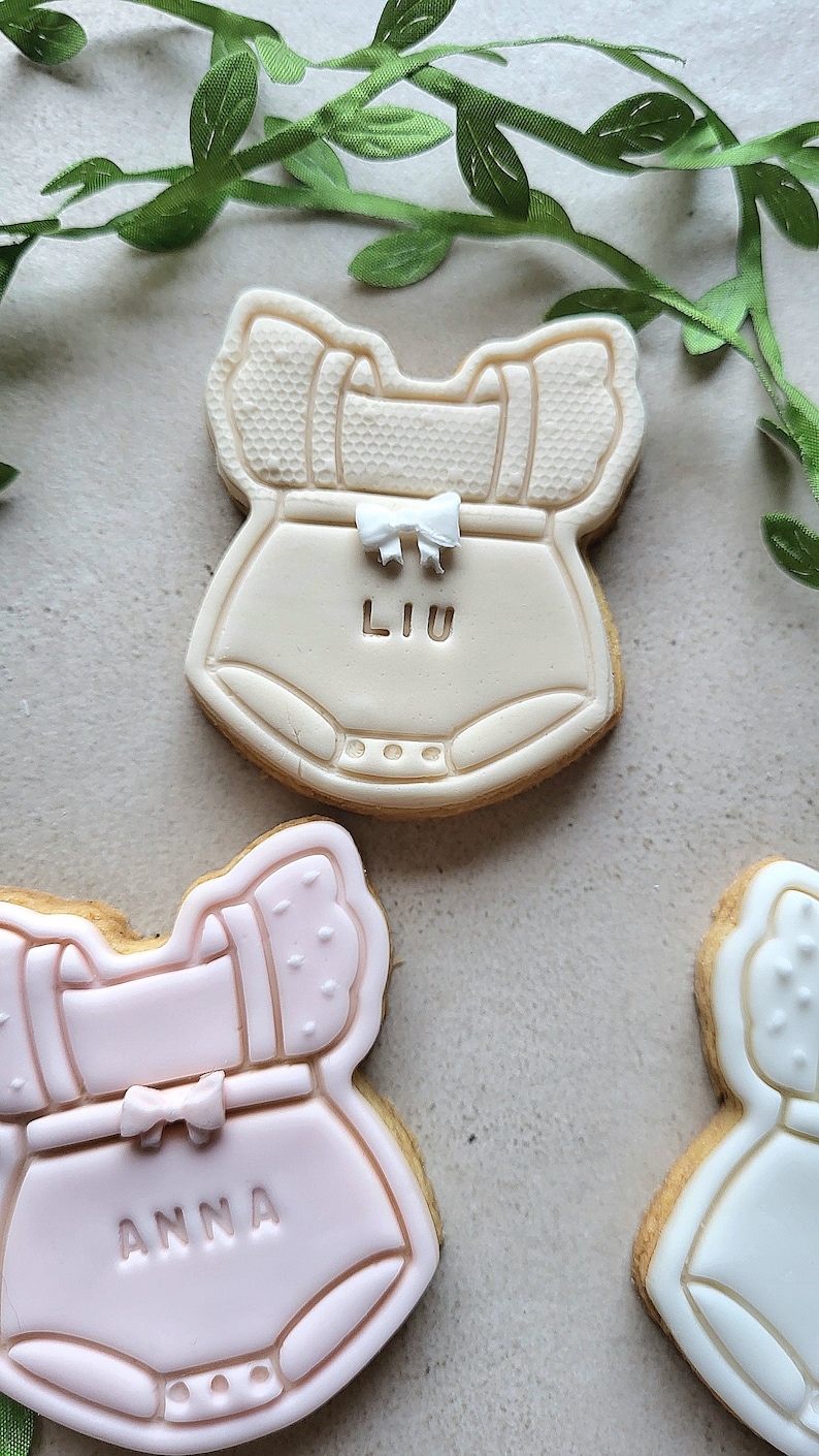 May include: Three decorated sugar cookies shaped like baby onesies. The top cookie is beige with a white bow and the name "LIU" written on it. The middle cookie is pink with a white bow and the name "ANNA" written on it. The bottom cookie is white with a white bow.