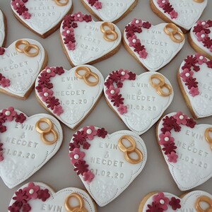 May include: Heart-shaped cookies decorated with white icing, gold rings, and burgundy flowers. The cookies are personalized with the text "EVIN & NECDET 01.02.20".