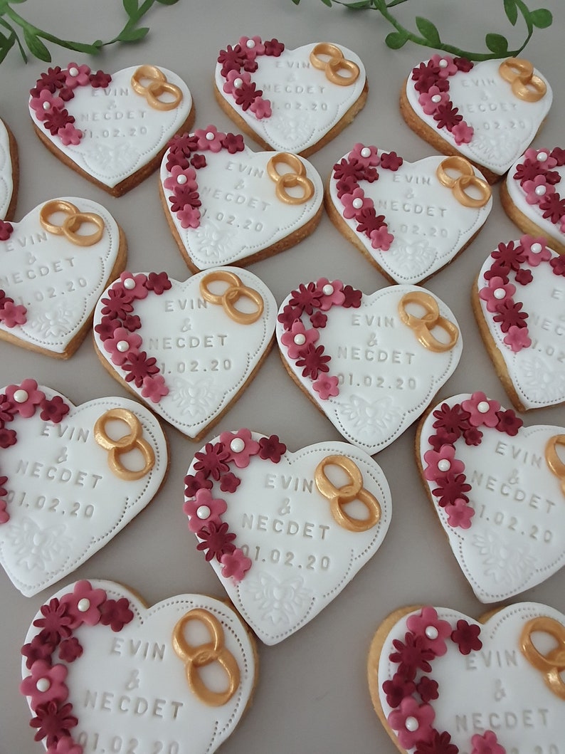 May include: Heart-shaped cookies decorated with white icing, gold rings, and burgundy flowers. The cookies are personalized with the text "EVIN & NECDET 01.02.20".