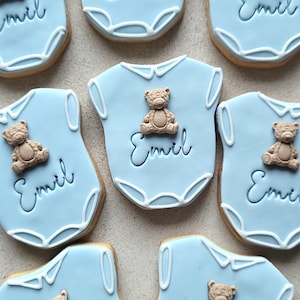 May include: Light blue baby onesie cookies decorated with a teddy bear and the name "Emil" in cursive. The cookies have a white trim and are arranged on a light-colored surface. These cookies are perfect for a baby shower or a newborn celebration.