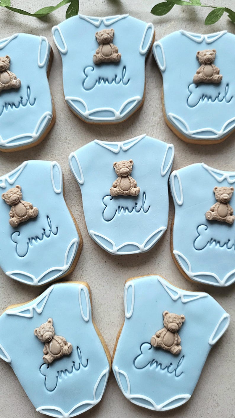May include: Light blue baby onesie cookies decorated with a teddy bear and the name "Emil" in cursive. The cookies have a white trim and are arranged on a light-colored surface. These cookies are perfect for a baby shower or a newborn celebration.