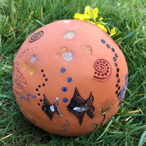 Könnte beinhalten: Eine Terrakotta-Kugel mit verschiedenen bemalten Designs, darunter Fische, Muscheln und Punkte. Die Kugel ist in den Farben Blau, Weiß, Gelb und Schwarz verziert.
