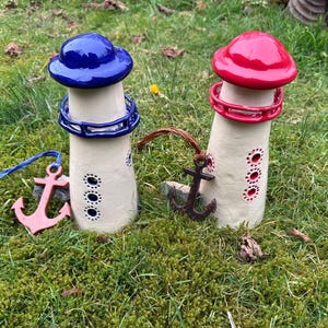 Könnte beinhalten: Zwei Keramik-Leuchtturm-Figuren mit blauen und roten Dächern, die auf einem Bett aus grünem Moos stehen. Jeder Leuchtturm hat einen kleinen Anker-Anhänger.
