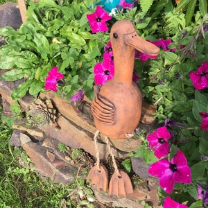 Könnte beinhalten: Eine Terrakotta-Entenfigur mit langem Schnabel und Schnüren als Beinen. Die Ente sitzt auf einer Steinmauer mit grünen Pflanzen und rosa Blumen im Hintergrund.