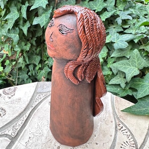 Könnte beinhalten: Eine terrakottafarbene Keramikskulptur eines menschlichen Kopfes und Oberkörpers. Die Figur zeigt geschnitztes Haar mit strukturierten Strähnen, geschlossene Augen und eine definierte Nase. Die Oberfläche hat ein mattes, erdiges Finish.