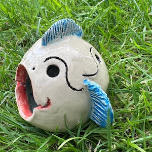 Könnte beinhalten: Eine weiße Keramikfischskulptur mit blauen und roten Akzenten. Der Fisch hat einen großen, offenen Mund und ein schwarzes Auge. Die Skulptur steht auf einem Bett aus grünem Gras.