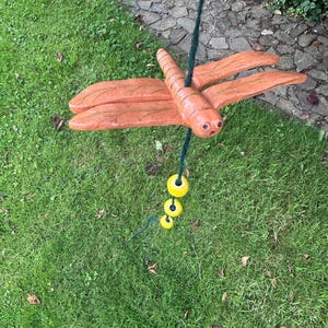 Könnte beinhalten: Eine handgefertigte orangefarbene Libellen-Schaukel, die an einem grünen Seil hängt. Die Libelle hat detaillierte Flügel und einen segmentierten Körper. Drei gelbe Kugeln sind unten am Seil befestigt. Die Schaukel befindet sich vor dem Hintergrund von grünem Gras und einem Steinweg.