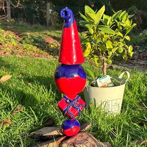 Könnte beinhalten: Eine Keramik-Garten-Zwergen-Dekoration mit einem rot-blauen Hut, einer rot-blauen Kugel, einer rot-blauen Geschenkbox und einer rot-blauen Kugel. Der Zwerg steht auf einem Holzstamm in einem grasbewachsenen Bereich.