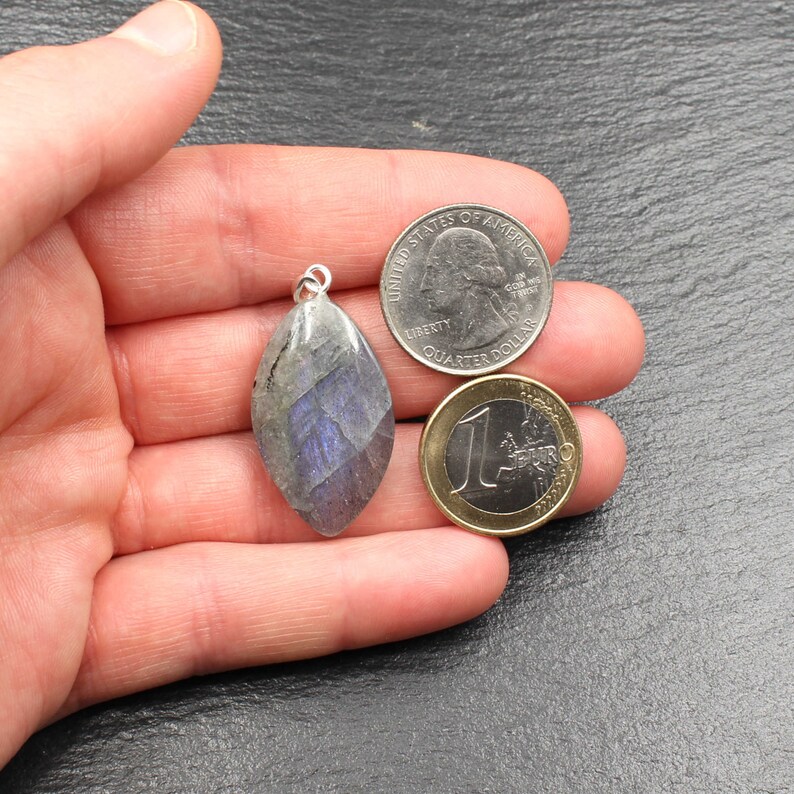 Op de afbeelding: Een gefacetteerde, druppelvormige labradoriet hanger met een zilveren balein. De hanger toont iriserende blauwe en grijze tinten. Een Amerikaanse kwart dollar en een euromunt liggen naast de hanger ter vergelijking.