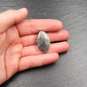Op de afbeelding: Een gepolijste, druppelvormige hanger met een zilveren beugel. De steen heeft een mix van grijze, groene en zilveren tinten, met een subtiel, natuurlijk patroon. De hanger wordt in een hand gehouden tegen een donkere achtergrond, wat de unieke kleur en vorm benadrukt.
