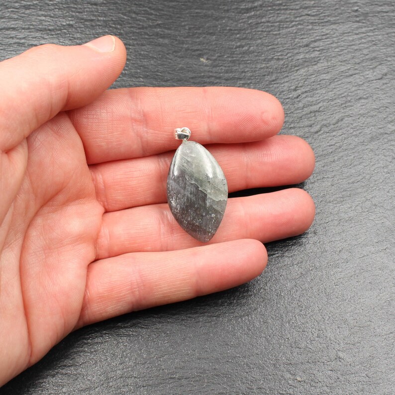 Op de afbeelding: Een gepolijste, druppelvormige hanger met een zilveren beugel. De steen heeft een mix van grijze, groene en zilveren tinten, met een subtiel, natuurlijk patroon. De hanger wordt in een hand gehouden tegen een donkere achtergrond, wat de unieke kleur en vorm benadrukt.
