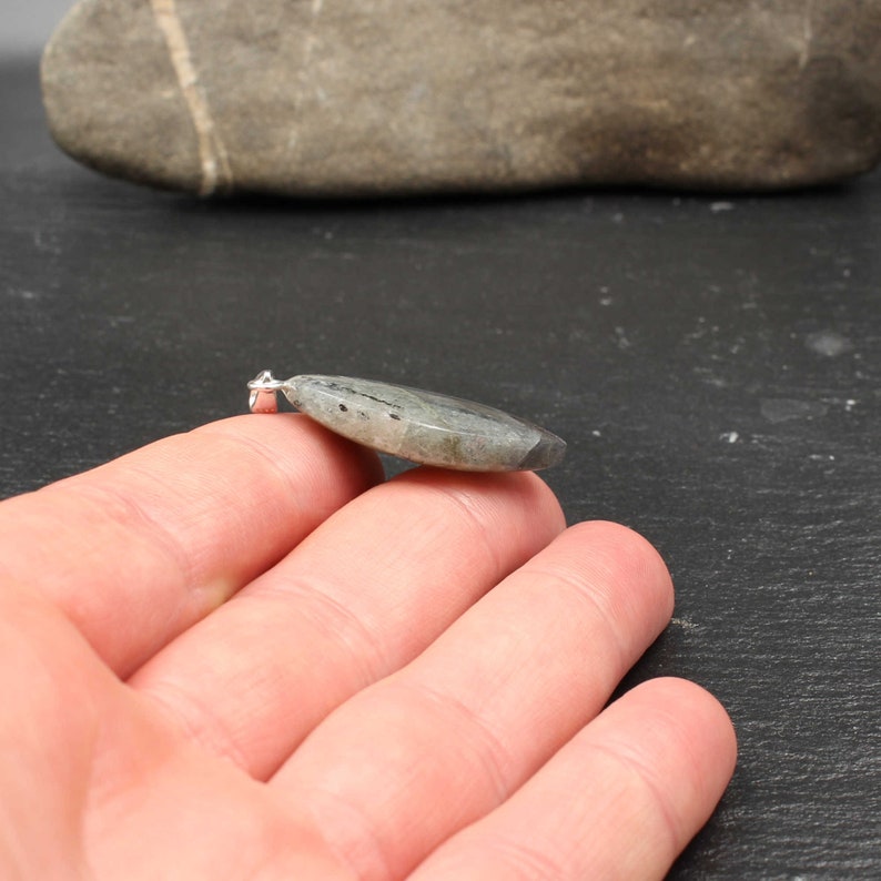 Op de afbeelding: Een ovale hanger van grijsgroene steen met een zilverkleurige balein. De hanger heeft natuurlijke patronen en kleurvariaties. De gepolijste steen is ongeveer 4 cm lang en klaar om aan een ketting te worden gedragen.