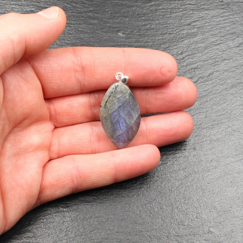 Op de afbeelding: Een gefacetteerde, druppelvormige hanger met een zilveren beugel. De edelsteen vertoont een mix van grijze, blauwe en paarse tinten. De hanger wordt in een hand gehouden tegen een donkergrijze achtergrond. De edelsteen is ongeveer 3,8 cm lang.