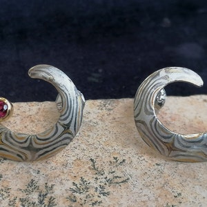 Könnte beinhalten: Ein Paar Ohrringe aus Silber und Gold mit einem wirbelnden Muster und einem rosafarbenen Edelstein-Akzent.