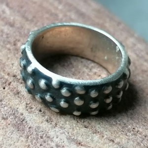 Op de afbeelding: Een zilveren ring met een textuurontwerp. De ring heeft een band van kleine, gelijkmatig verdeelde, verhoogde cirkelvormige details. Het metaal heeft een licht donkerder uiterlijk, waardoor een contrast ontstaat met de helderdere, gepolijste oppervlakken van de ring.