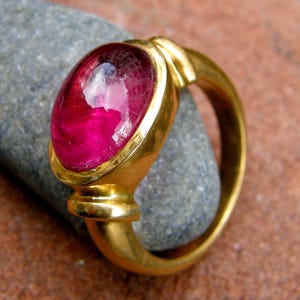 Könnte beinhalten: Ein goldener Ring mit einem großen, oval geformten, rubinroten Stein. Band und Fassung des Rings sind aus Gold. Der Stein hat eine leuchtende, rötlich-rosa Farbe. Der Ring liegt auf einem grauen Stein, der das Design und die Farbe des Schmucks hervorhebt.