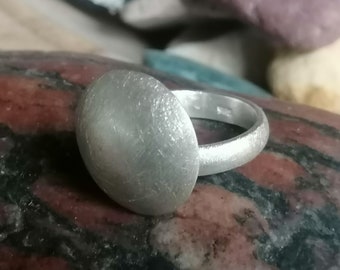 May include: A silver ring with a large, round, domed top. The ring has a brushed finish and is set on a textured, red and brown surface.
