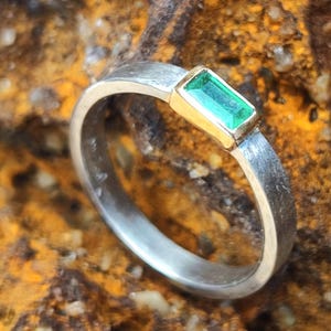 Può includere: Anello in argento con una gemma verde rettangolare taglio smeraldo incastonata in una lunetta dorata. L'anello ha una finitura in metallo spazzolato. Sfondo marrone strutturato.