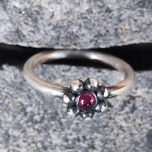 Può includere: Un anello d'argento con castone a forma di fiore. Il fiore ha una gemma rossa, rotonda e centrale, circondata da piccoli petali d'argento. La fascia dell'anello è liscia e argentata.