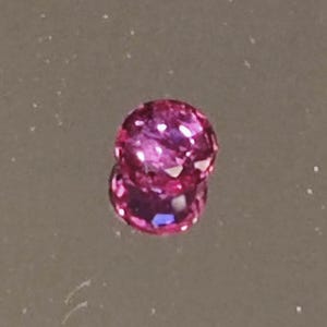 Könnte beinhalten: Ein runder, facettierter, magentafarbener Edelstein mit Brillantschliff. Der Edelstein liegt auf einer reflektierenden Oberfläche, wodurch ein Spiegeleffekt entsteht. Die Facetten des Edelsteins fangen das Licht ein und zeigen eine Reihe von rosa und violetten Farbtönen.