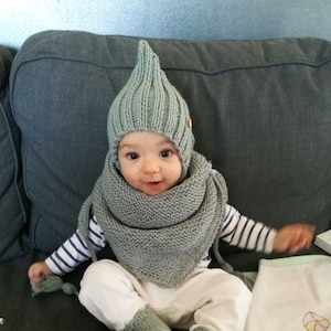 Può includere: Un bambino che indossa un cappello di lana verde chiaro con la cima a punta e sciarpa e scarpine abbinate. Il bambino è seduto su un divano grigio e sorride.