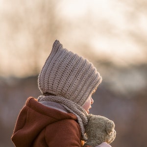Può includere: Un bambino indossa una giacca marrone con cappuccio e un cappello di lana beige e tiene un orsacchiotto di peluche marrone. Il bambino è seduto all'aperto con uno sfondo sfocato.