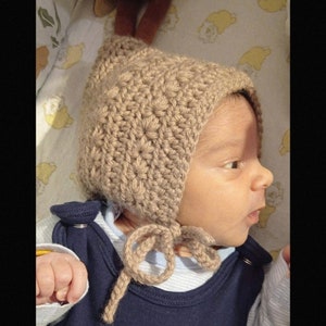 Può includere: Un bambino che indossa un berretto di lana marrone con un fiocco. Il berretto ha una punta a punta e copre le orecchie del bambino.
