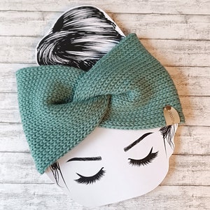 Könnte beinhalten: Ein grünes Strick Stirnband mit einem geknoteten Design. Das Stirnband hat ein kleines braunes Etikett mit dem Wort "Wollherz" darauf.