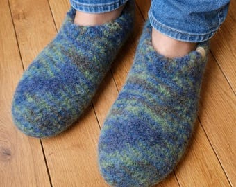 Könnte beinhalten: Ein Paar blau-grüne Wollpantoffeln. Die Pantoffeln haben ein strukturiertes, gesprenkeltes Aussehen und werden mit einer blauen Jeans getragen. Die Pantoffeln liegen auf einem Holzboden.