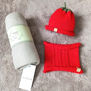 Könnte beinhalten: Eine gerollte hellgraue Strickdecke mit einer Papierbanderole mit der Aufschrift "kocyk" und "bamboo", eine rote Strickmütze mit grünem Stiel oben und ein roter Strickschal. Ein weißes Etikett mit dem Text "ANDER" ist an der Decke befestigt.