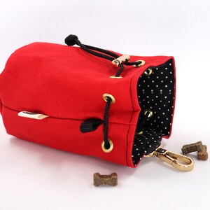 May include: A red canvas dog treat bag with a black drawstring closure and gold-colored hardware. The interior features a black and white polka dot pattern. Several dog treats are visible near the bag.