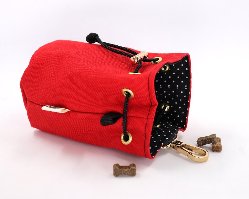 May include: A red canvas dog treat bag with a black drawstring closure and gold-colored hardware. The interior features a black and white polka dot pattern. Several dog treats are visible near the bag.