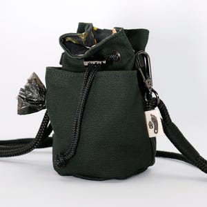 Op de afbeelding: Een donkergroene canvas tas met een trekkoord en een zwarte band. De tas heeft een klein vakje aan de voorkant en een zwarte karabijnhaak die aan de band is bevestigd. Een kleine plastic zak is aan de band bevestigd.