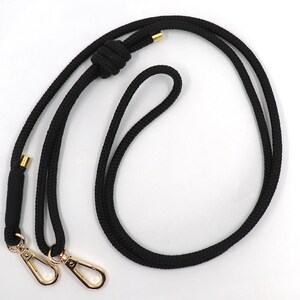 May include: Black rope lanyard with gold-colored metal swivel clasps and accents. The lanyard features a knotted design and is suitable for holding keys, an ID badge, or other small items. The rope is thick and durable.