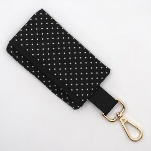 May include: Black key holder with white polka dots. The key holder has a black strap and a gold-colored metal clasp. The key holder is folded over.