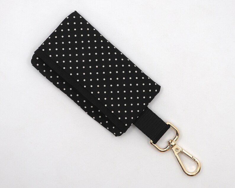 May include: Black key holder with white polka dots. The key holder has a black strap and a gold-colored metal clasp. The key holder is folded over.