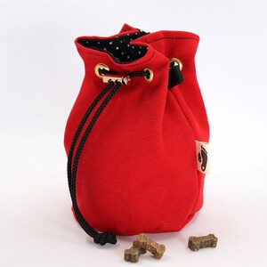 May include: A bright red drawstring pouch with a black drawstring closure and gold-colored grommets. The interior lining is black with white polka dots. A small tag with a footprint logo is attached to the side. Several dog treats are in front of the pouch.