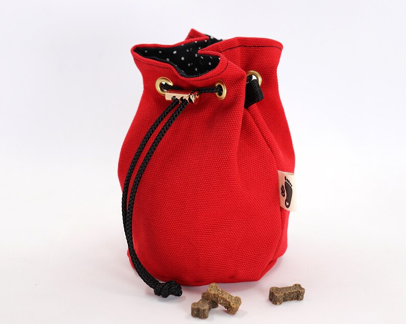 May include: A bright red drawstring pouch with a black drawstring closure and gold-colored grommets. The interior lining is black with white polka dots. A small tag with a footprint logo is attached to the side. Several dog treats are in front of the pouch.