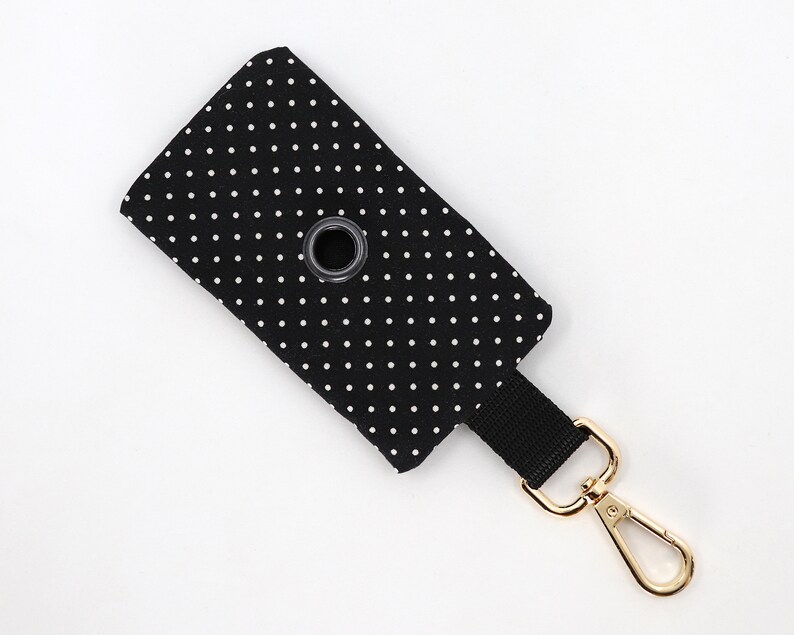 May include: Black and white polka dot waste bag dispenser with a black strap and gold-colored clasp. The dispenser has a metal grommet for easy bag access. A stylish accessory for pet owners.