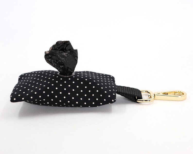 May include: A black and white polka dot dog waste bag dispenser with a black strap and gold-colored clasp. A roll of black waste bags is visible, dispensing from the top of the dispenser. The dispenser is designed for pet owners.