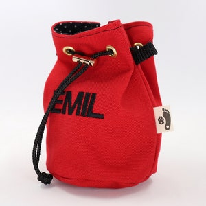 May include: A red canvas drawstring bag with the name "EMIL" embroidered in black. The bag has a black drawstring, gold-colored grommets, and a small tag with a paw print and heart logo. The interior lining is black with white polka dots.