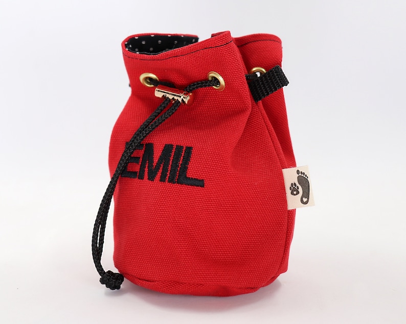 May include: A red canvas drawstring bag with the name "EMIL" embroidered in black. The bag has a black drawstring, gold-colored grommets, and a small tag with a paw print and heart logo. The interior lining is black with white polka dots.