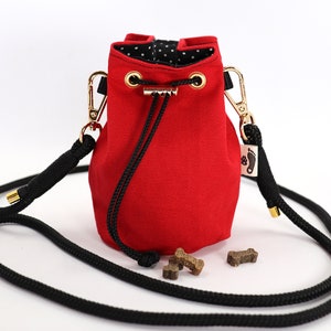 May include: A red canvas dog treat bag with a black rope strap and gold-colored hardware. The bag has a drawstring closure and a black and white polka dot lining. Several dog treats are visible near the bag.
