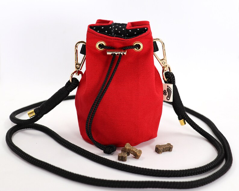 May include: A red canvas dog treat bag with a black rope strap and gold-colored hardware. The bag has a drawstring closure and a black and white polka dot lining. Several dog treats are visible near the bag.