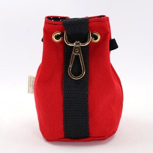 May include: A small, red canvas drawstring bag with a black vertical stripe and a gold-colored clasp. The bag has a black strap and gold eyelets. The interior lining is white with black polka dots. A small tag is attached to the side.
