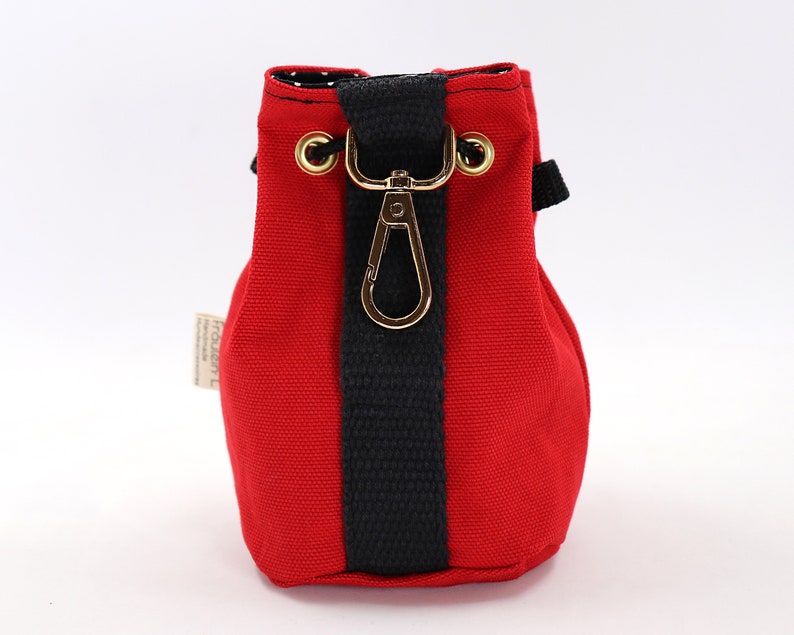 May include: A small, red canvas drawstring bag with a black vertical stripe and a gold-colored clasp. The bag has a black strap and gold eyelets. The interior lining is white with black polka dots. A small tag is attached to the side.