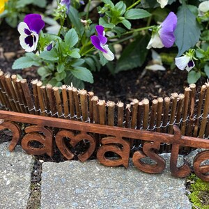 May include: A wooden sign with the word "namaste" in a stylized font, set against a backdrop of a small wooden fence and vibrant purple and white flowers. The sign is a warm brown color.