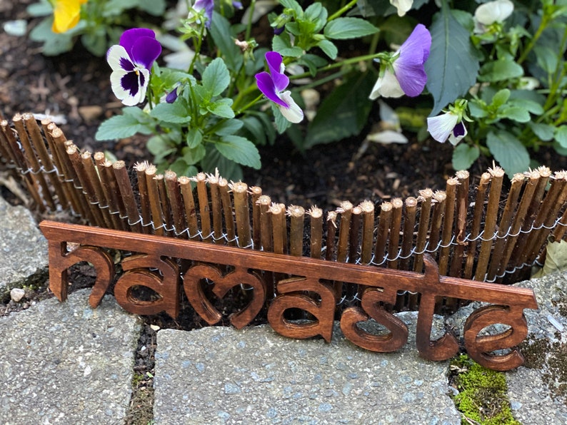 May include: A wooden sign with the word "namaste" in a stylized font, set against a backdrop of a small wooden fence and vibrant purple and white flowers. The sign is a warm brown color.