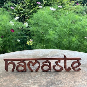 May include: A wooden sign with the word "namaste" in a stylized font. The sign is a dark brown color and sits on a stone surface, with a lush green garden and flowers in the background.
